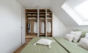 Een open kast met kleding en beddengoed, naast een groen opgemaakt bed onder een dakraam.