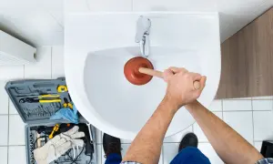Handen gebruiken een ontstopper om het wasbak putje schoonmaken. Gereedschapskist met tangen, boren en handschoenen ligt ernaast.