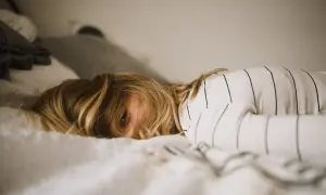 Een kind met blond haar ligt in bed, met een oog dat net zichtbaar is. De kamer is rommelig, het kind kan niet opruimen.