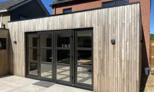 Moderne uitbouw voorkant huis met houten gevelbekleding en grote glazen deuren. De zon werpt schaduwen op de lichte terrastegels.