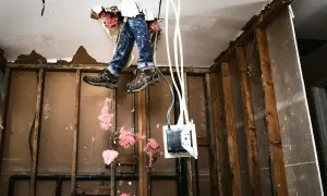 Man met werklaarzen en jeans hangt door een gat in het plafond, omringd door isolatie. Een doe-het-zelf project om zelf te verbouwen.