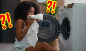 Vrouw ruikt aan een handdoek bij een wasmachine. Vraagt zich af hoe vaak handdoek gebruiken? Grote rode vraagtekens suggereren twijfel.