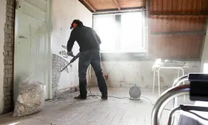Man met boorhamer sloopt muur voor goedkoop verbouwen. Stof dwarrelt in het zonlicht.
