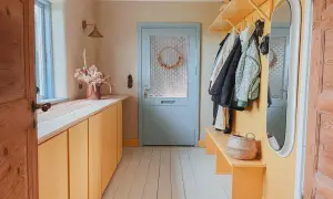 Gerenoveerde gang met gele kasten, kapstok en spiegel. Een mandje en droogbloemen maken het gezellig.