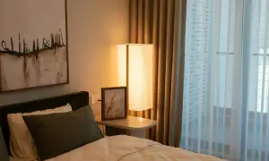 Een comfortabel bed voor een raam met lichte gordijnen. Een staande lamp en een schilderij aan de muur creëren een rustige sfeer.
