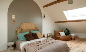 Een rustige, minimalistische slaapkamer met een rieten hoofdeinde, zachte plaids en een comfortabele fauteuil onder een dakraam.
