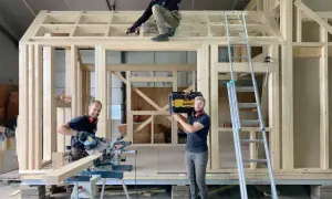 Drie mannen werken aan een **tiny huis bouwen**. Eén zaagt hout, een ander schroeft aan het dak, en de derde draagt een gereedschapskist.