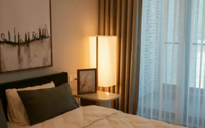 Een comfortabel bed voor een raam met lichte gordijnen. Een staande lamp en een schilderij aan de muur creëren een rustige sfeer.