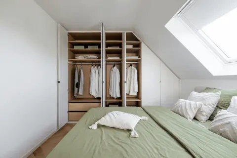 Een open kast met kleding en beddengoed, naast een groen opgemaakt bed onder een dakraam.