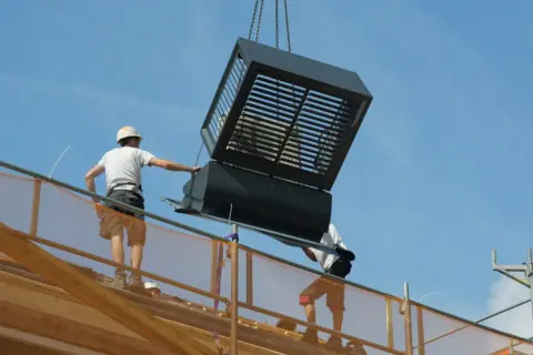 Bouwvakkers plaatsen een grote, prefab ventilatie-unit op een dak. Dit laat zien hoe wat is prefab: kant-en-klare onderdelen die snel gemonteerd worden.