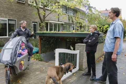 Een vader met dochter in bakfiets, zoon en hond staan buiten. Een slimme oplossing voor fietsen in de voortuin, met een groen dak boven de stalling.