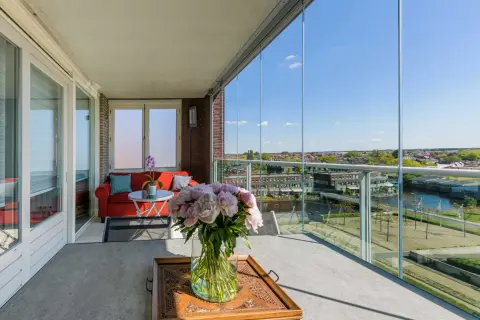 Een prachtig uitzicht vanaf dit balkon, dat met glazen schuifwanden is dichtgemaakt. Een rode bank en bloemen maken het gezellig.