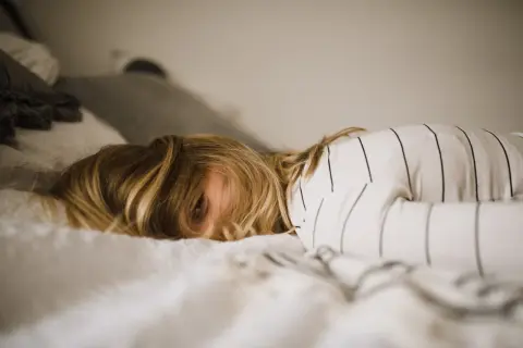 Een kind met blond haar ligt in bed, met een oog dat net zichtbaar is. De kamer is rommelig, het kind kan niet opruimen.