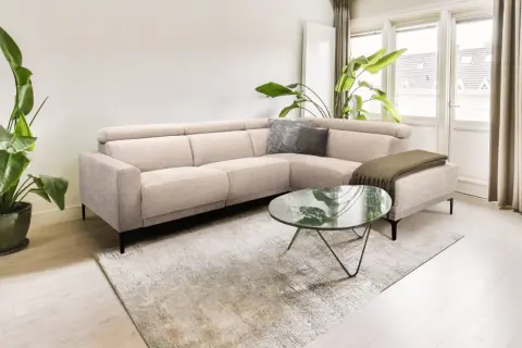 Licht beige hoekbank met kussens en plaid, een marmeren salontafel en grote planten creëren een rustig slow living interieur.