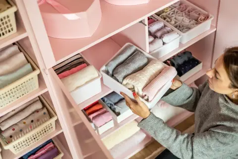 Vrouw sorteert netjes opgevouwen kleding in een roze kledingkast. Een georganiseerde kledingkast opruimen geeft rust.