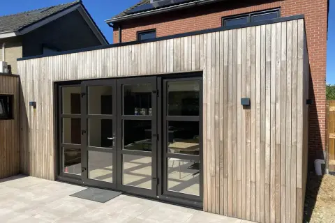 Moderne uitbouw voorkant huis met houten gevelbekleding en grote glazen deuren. De zon werpt schaduwen op de lichte terrastegels.
