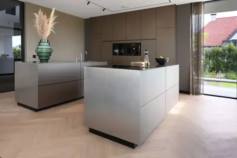 Moderne keuken met strakke, metalen kasten op een visgraat houten vloer. Een groene vaas met pampasgras siert het aanrecht.