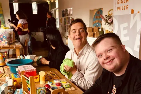 Twee lachende mannen met het syndroom van Down, omringd door speelgoed. Ze lijken klaar om het speelgoed op te ruimen, een gezellige activiteit.