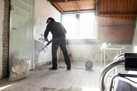 Man met boorhamer sloopt muur voor goedkoop verbouwen. Stof dwarrelt in het zonlicht.