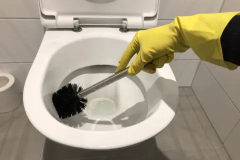 Hand met gele handschoen is bezig met wc poetsen. De toiletborstel maakt de pot schoon.