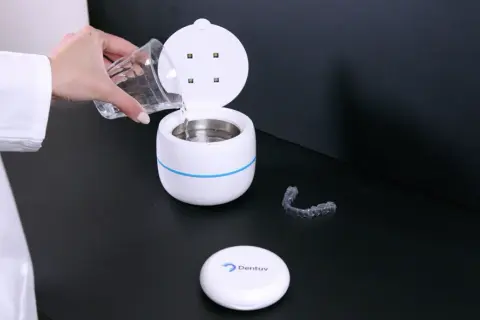 Een hand met een glas water vult een apparaat voor ultrasoon reinigen. Een bitje ligt ernaast.