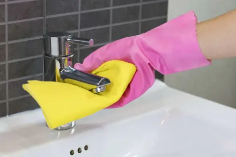 Hand in roze handschoen maakt de kraan van de wasbak schoon met een gele doek.