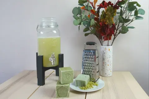 Zelf wasmiddel maken ervaringen: groene zeepblokken, rasp, en een glazen dispenser met gele vloeistof. Een vaas met herfstbladeren completeert het tafereel.