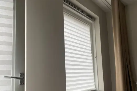 Wit plisségordijn als tijdelijke raambekleding tijdens een verbouwing. Een beige gordijn hangt ernaast.