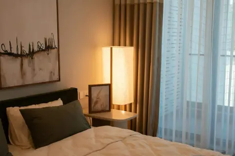 Een comfortabel bed voor een raam met lichte gordijnen. Een staande lamp en een schilderij aan de muur creëren een rustige sfeer.