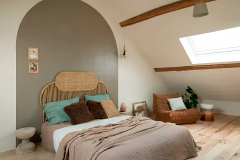 Een rustige, minimalistische slaapkamer met een rieten hoofdeinde, zachte plaids en een comfortabele fauteuil onder een dakraam.