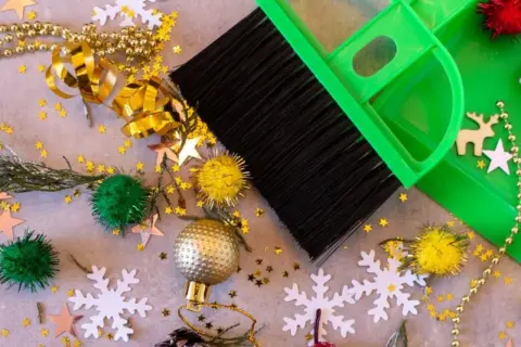 Een groene bezem en stoffer maken de kerstspullen op, met glitters, kerstballen en sneeuwvlokken verspreid.