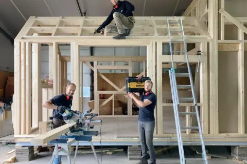 Drie mannen werken aan een **tiny huis bouwen**. Eén zaagt hout, een ander schroeft aan het dak, en de derde draagt een gereedschapskist.