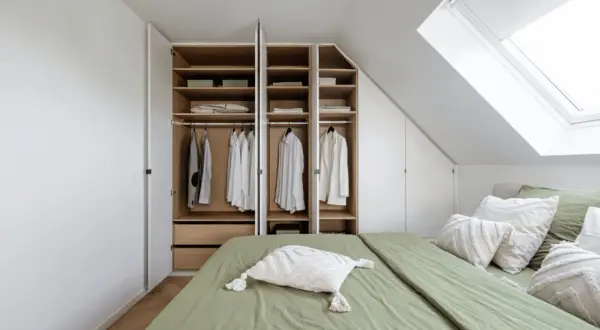 Een open kast met kleding en beddengoed, naast een groen opgemaakt bed onder een dakraam.