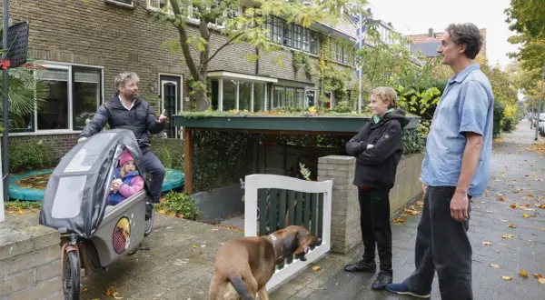 Een vader met dochter in bakfiets, zoon en hond staan buiten. Een slimme oplossing voor fietsen in de voortuin, met een groen dak boven de stalling.