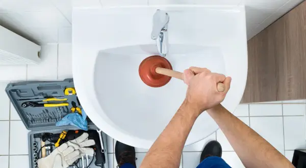 Handen gebruiken een ontstopper om het wasbak putje schoonmaken. Gereedschapskist met tangen, boren en handschoenen ligt ernaast.