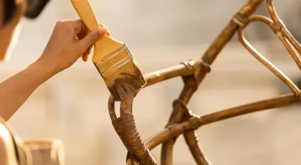 Hand met kwast werkt aan het **zelf rotan stoel repareren**. De stoel krijgt een opknapbeurt.