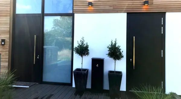 Moderne zwarte voordeur met gouden handgreep, naast een groot raam. Twee olijfbomen in potten en een zwarte brievenbus met nummer 4 bieden voordeur inspiratie.
