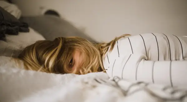 Een kind met blond haar ligt in bed, met een oog dat net zichtbaar is. De kamer is rommelig, het kind kan niet opruimen.
