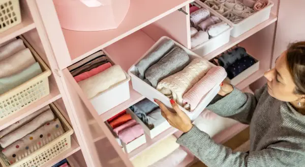 Vrouw sorteert netjes opgevouwen kleding in een roze kledingkast. Een georganiseerde kledingkast opruimen geeft rust.