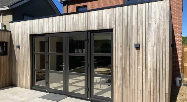 Moderne uitbouw voorkant huis met houten gevelbekleding en grote glazen deuren. De zon werpt schaduwen op de lichte terrastegels.