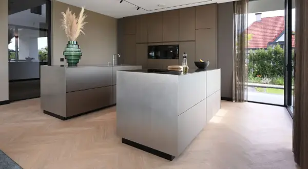 Moderne keuken met strakke, metalen kasten op een visgraat houten vloer. Een groene vaas met pampasgras siert het aanrecht.
