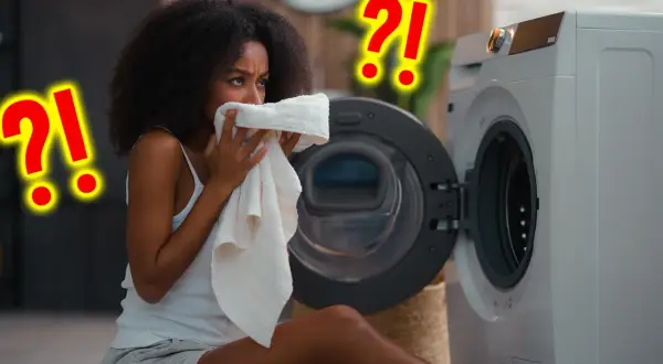 Vrouw ruikt aan een handdoek bij een wasmachine. Vraagt zich af hoe vaak handdoek gebruiken? Grote rode vraagtekens suggereren twijfel.