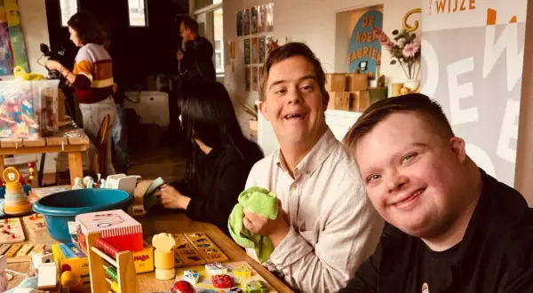 Twee lachende mannen met het syndroom van Down, omringd door speelgoed. Ze lijken klaar om het speelgoed op te ruimen, een gezellige activiteit.
