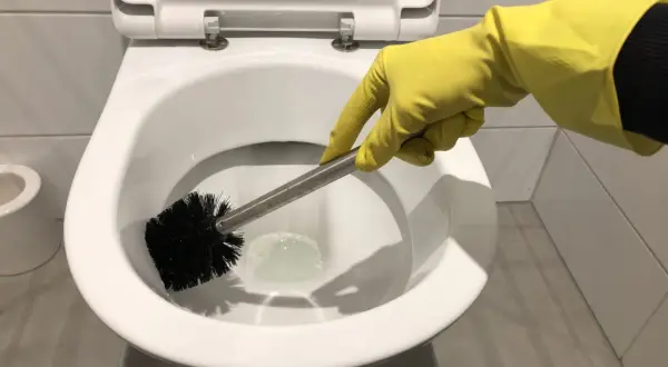 Hand met gele handschoen is bezig met wc poetsen. De toiletborstel maakt de pot schoon.