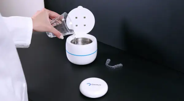 Een hand met een glas water vult een apparaat voor ultrasoon reinigen. Een bitje ligt ernaast.