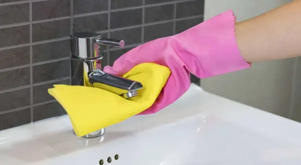 Hand in roze handschoen maakt de kraan van de wasbak schoon met een gele doek.