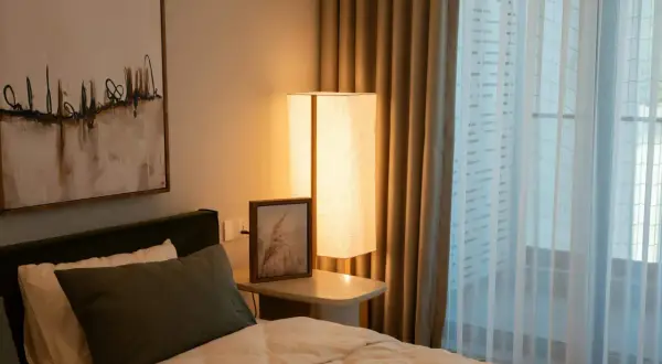 Een comfortabel bed voor een raam met lichte gordijnen. Een staande lamp en een schilderij aan de muur creëren een rustige sfeer.