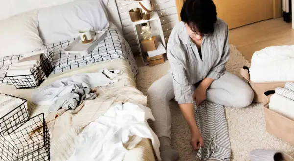 Een vrouw is bezig met het kamer opruimen. Ze vouwt kleding op en legt deze in manden naast een opgemaakt bed.
