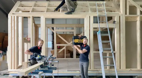 Drie mannen werken aan een **tiny huis bouwen**. Eén zaagt hout, een ander schroeft aan het dak, en de derde draagt een gereedschapskist.