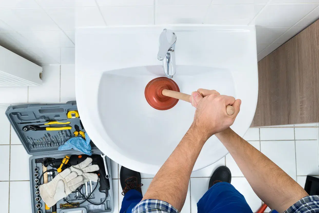 Handen gebruiken een ontstopper om het wasbak putje schoonmaken. Gereedschapskist met tangen, boren en handschoenen ligt ernaast.
